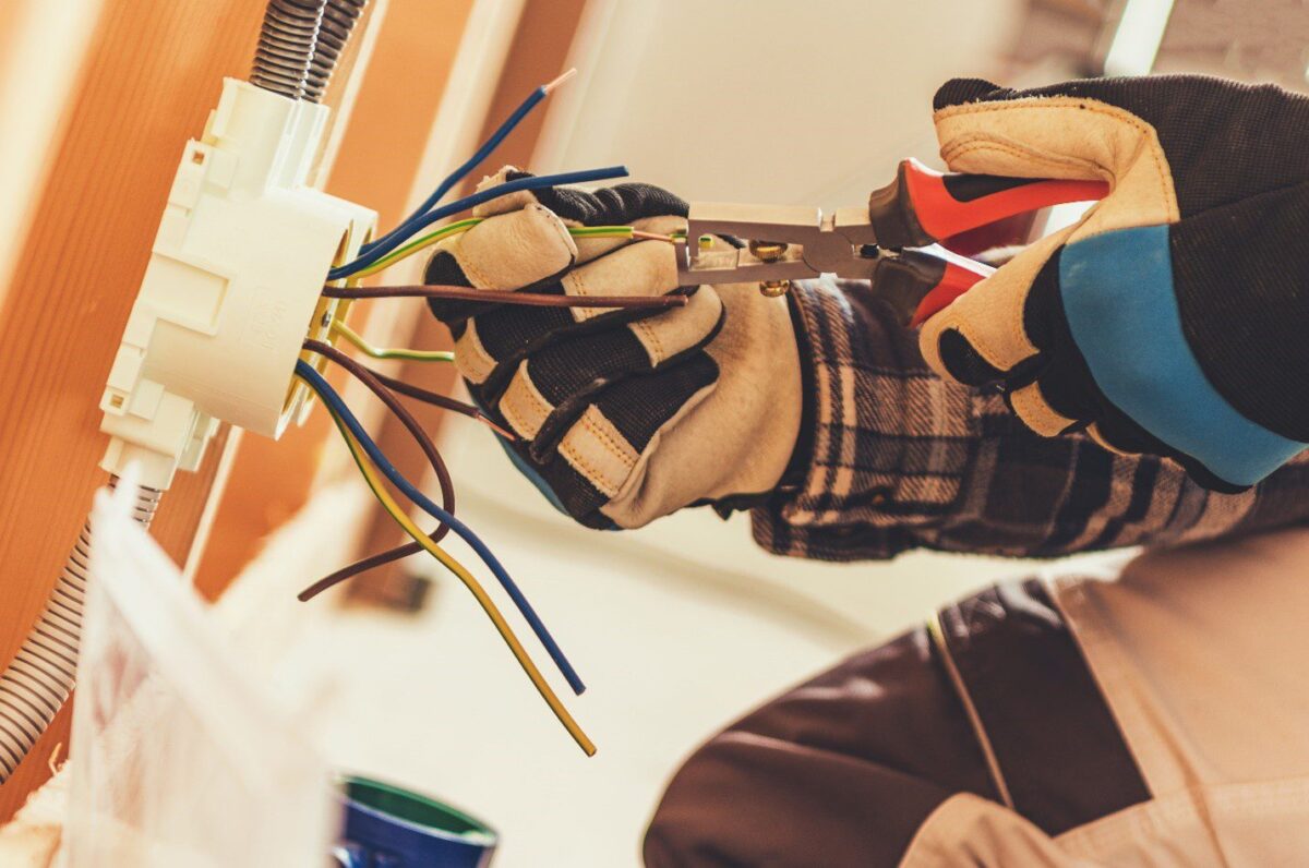 Electrician wearing gloves uses pliers to work on multi-colored wires protruding from a wall socket.