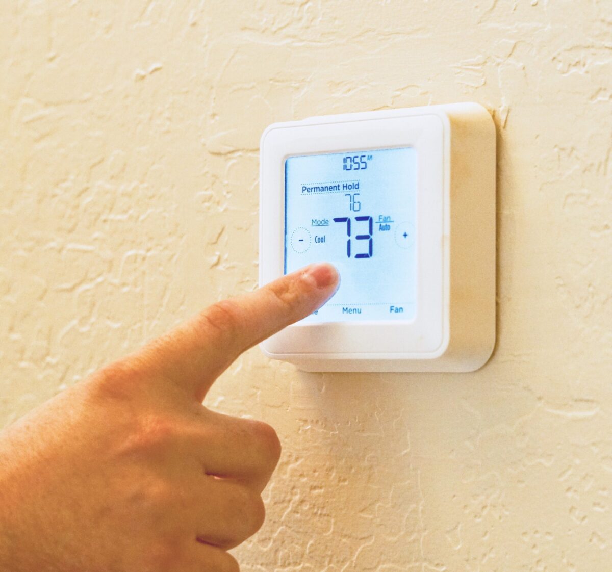A hand adjusts a white digital thermostat on a textured beige wall displaying 73 degrees.