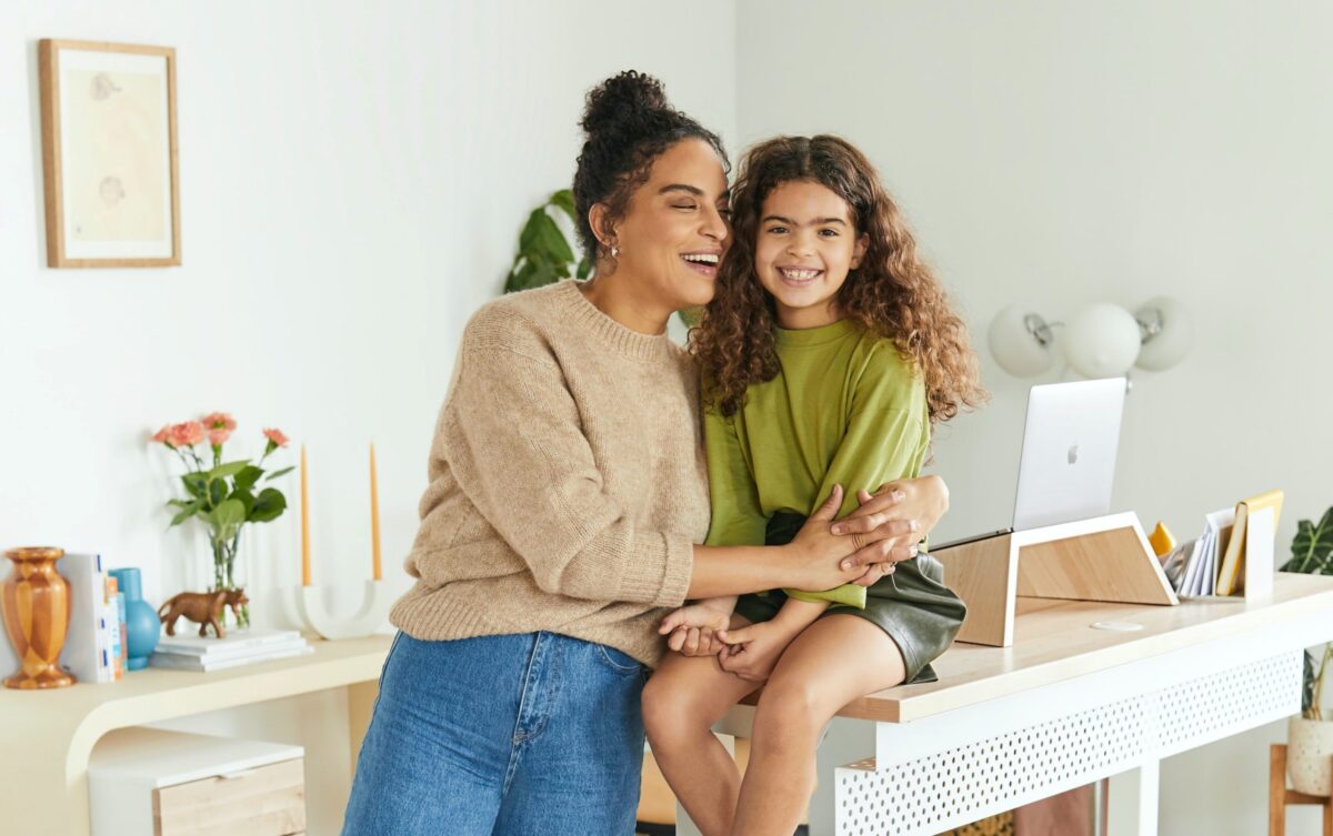 A woman and a girl smile, sitting on a desk in a room with plants and a laptop.