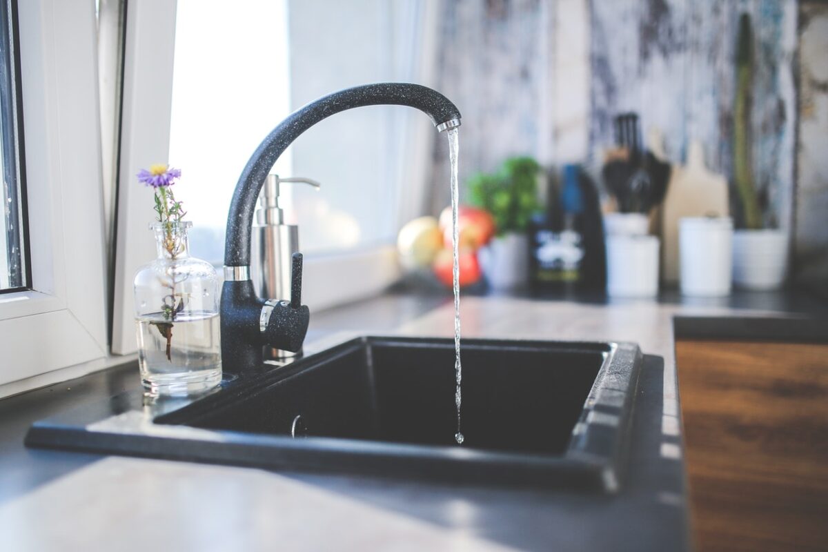 Water flows from a black kitchen faucet into a black sink. A small glass vase with flowers is next to the sink.