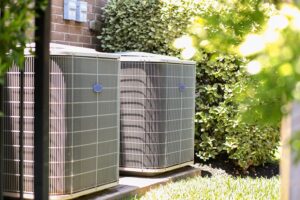 Two outdoor air conditioning units are installed beside a brick building, surrounded by green shrubs and grass.
