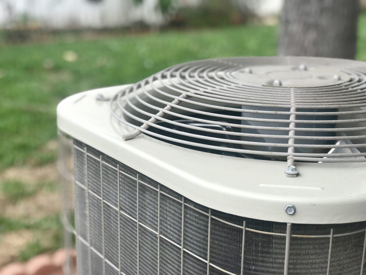 Close-up of an outdoor air conditioning unit with metal grilles and fan, set in a grassy yard.