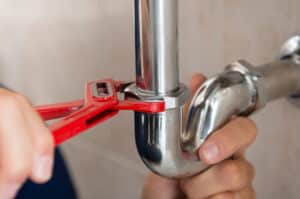 A person tightens a metal pipe with a red wrench, focusing on connecting a shiny U-bend joint.