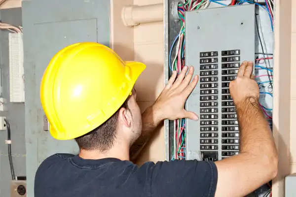 A Person In A Yellow Hard Hat Expertly Adjusts Wires Inside A Large Electrical Panel, Focusing On Circuit Breaker Repair.