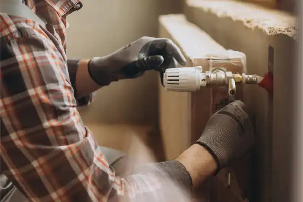Person in plaid shirt and gloves adjusting a valve on a radiator in a room, ensuring optimal performance like during a dehumidifier repair.