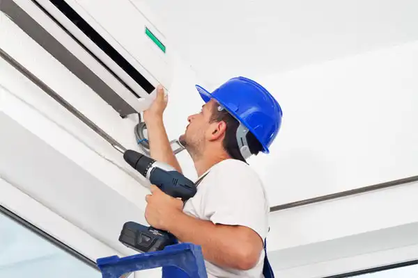 A Worker In A Blue Hard Hat On A Ladder Uses A Drill To Install Or Repair An Air Conditioning Unit Indoors.